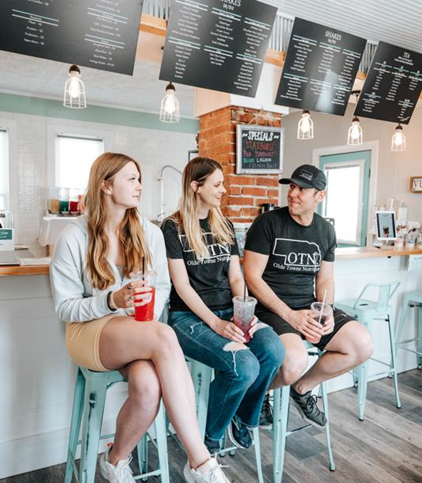 Three people sitting on teal stools at the Olde Towne Nutrition bar, chalkboard menus overhead